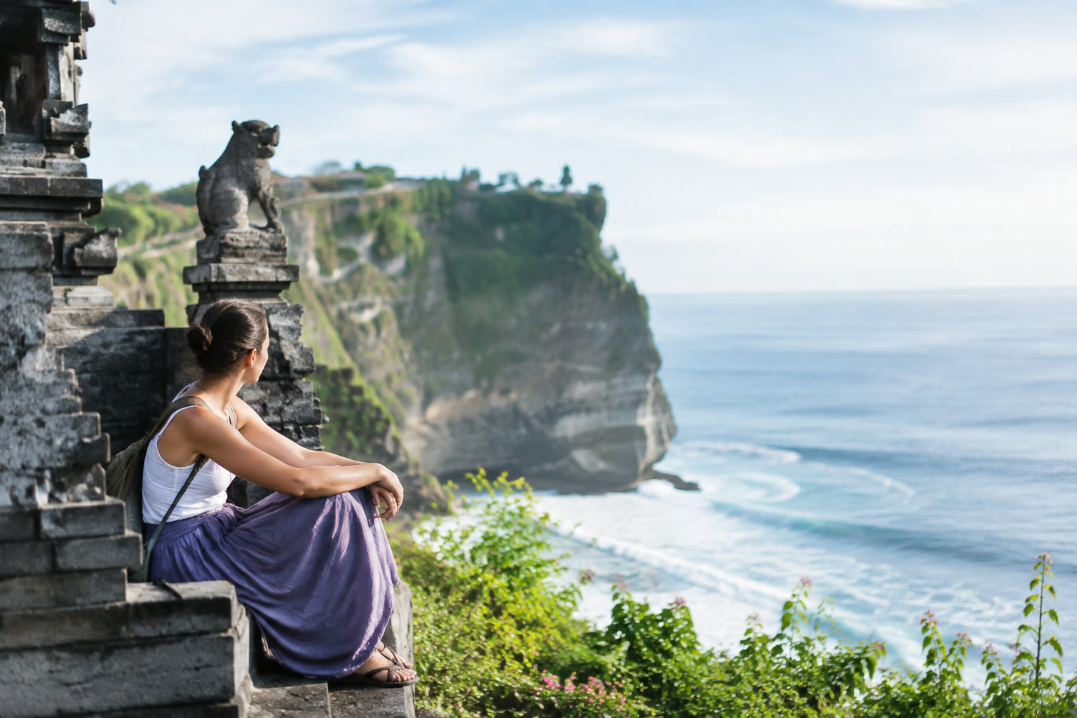 An anonymous traveller with an open notebook faces the sunrise on a quiet Bali beach, with outrigger fishing boats along the shore.